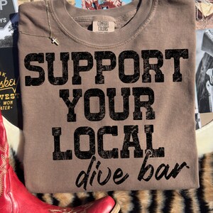 Peut inclure: Un sweat-shirt Comfort Colors marron avec le texte "SUPPORT YOUR LOCAL dive bar" en lettres noires délavées. Un collier argenté avec un pendentif en forme de croix est sur le sweat-shirt. Le sweat-shirt est sur une surface à motifs.