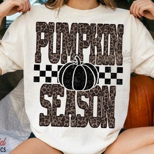 Può includere: Una t-shirt a maniche corte color crema con la scritta PUMPKIN SEASON in un motivo leopardato marrone. Tra le parole si trova una fascia a scacchi bianchi e neri e una grafica di una zucca nera al centro. Il design è centrato sul petto.