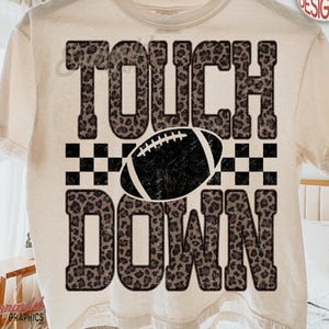 Könnte beinhalten: Ein cremefarbenes Kurzarm-T-Shirt mit dem Schriftzug TOUCH DOWN in einem braun-schwarzen Leopardenmuster. Eine schwarze Football-Grafik ist zwischen den Wörtern zentriert und wird von schwarz-weiß karierten Quadraten flankiert.
