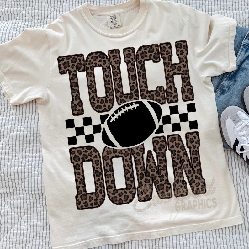 Puede incluir: Una camiseta de manga corta color crema con el texto TOUCH DOWN en una fuente con estampado de leopardo marr&oacute;n. Un gr&aacute;fico de bal&oacute;n de f&uacute;tbol americano en blanco y negro se sit&uacute;a entre las palabras, junto a un patr&oacute;n de cuadros blanco y negro.