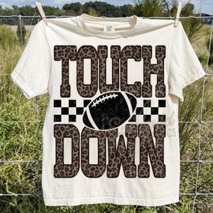 Puede incluir: Una camiseta de manga corta de color crema con el texto "TOUCH DOWN" en una fuente con estampado de leopardo marr&oacute;n. Entre las palabras hay un gr&aacute;fico de un bal&oacute;n de f&uacute;tbol americano en blanco y negro sobre un fondo de cuadros.
