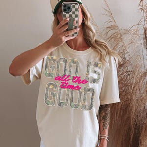 K&ouml;nnte beinhalten: Cremefarbenes T-Shirt mit dem Text "GOD IS all the time GOOD" in Tarn- und rosa Schrift. Die Person tr&auml;gt eine Baseballkappe mit Schachbrettmuster und h&auml;lt ein Telefon.