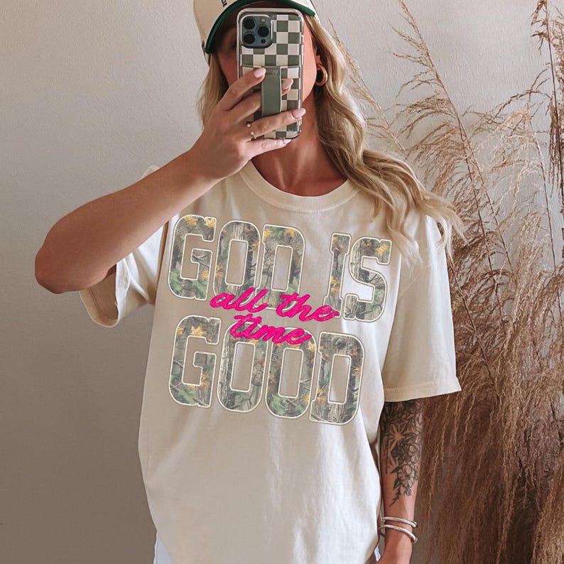 K&ouml;nnte beinhalten: Cremefarbenes T-Shirt mit dem Text "GOD IS all the time GOOD" in Tarn- und rosa Schrift. Die Person tr&auml;gt eine Baseballkappe mit Schachbrettmuster und h&auml;lt ein Telefon.