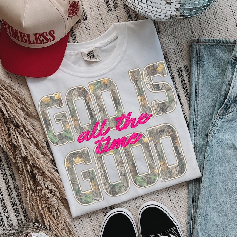 K&ouml;nnte beinhalten: Wei&szlig;es T-Shirt mit dem Text "GOD IS all the time GOOD" in einem Camouflage-Muster und rosa Stickerei. Eine rot-beige Baseballkappe mit dem Wort "TIMELESS" ist ebenfalls zu sehen.
