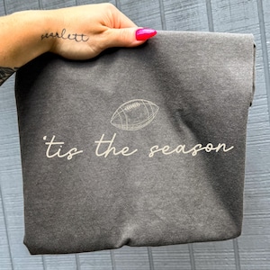 May include: A charcoal gray t-shirt featuring a white line-art illustration of an American football above the white cursive text 'tis the season'. The fabric has a slightly faded, vintage-inspired texture and a soft, casual appearance.