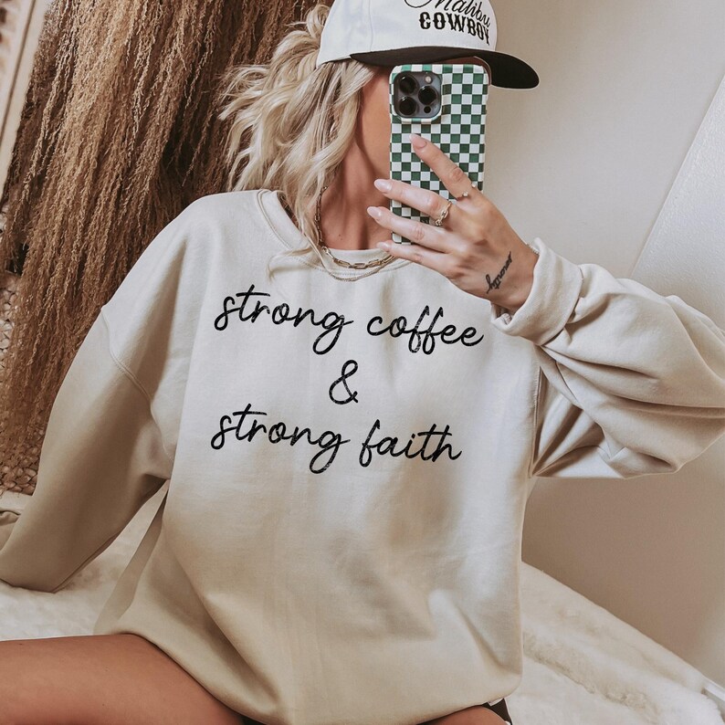 May include: Cream-colored sweatshirt with the text "strong coffee & strong faith" in black script. The sweatshirt has long sleeves and a crew neck. A white baseball cap with black text is visible.