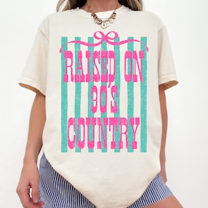 May include: Cream-colored t-shirt with the text "RAISED ON 90'S COUNTRY" in pink, set against a backdrop of teal vertical stripes. A pink bow is at the top of the design.