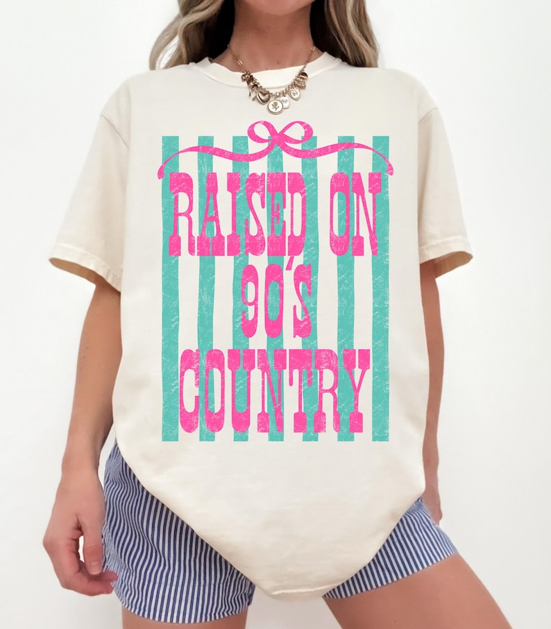 May include: Cream-colored t-shirt with the text "RAISED ON 90'S COUNTRY" in pink, set against a backdrop of teal vertical stripes. A pink bow is at the top of the design.