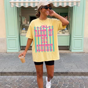 May include: A yellow t-shirt with the text "RAISED ON 90'S COUNTRY" in pink and teal. The shirt is worn with black shorts and white sneakers. The person is wearing sunglasses and a baseball cap.