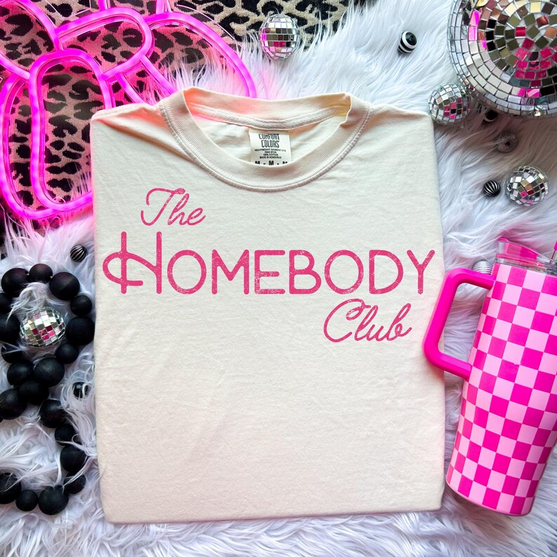 K&ouml;nnte beinhalten: Cremefarbenes T-Shirt mit dem pinkfarbenen Text "The Homebody Club". Das Shirt wird auf einer flauschigen wei&szlig;en Oberfl&auml;che pr&auml;sentiert, zusammen mit einem pinkfarbenen karierten Becher und Discokugel-Akzenten.