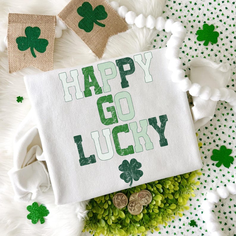 May include: A white sweatshirt with the words "HAPPY GO LUCKY" in green lettering and a shamrock graphic. The image includes decorative shamrock banners, coins, and a green plant, all on a white fluffy surface.