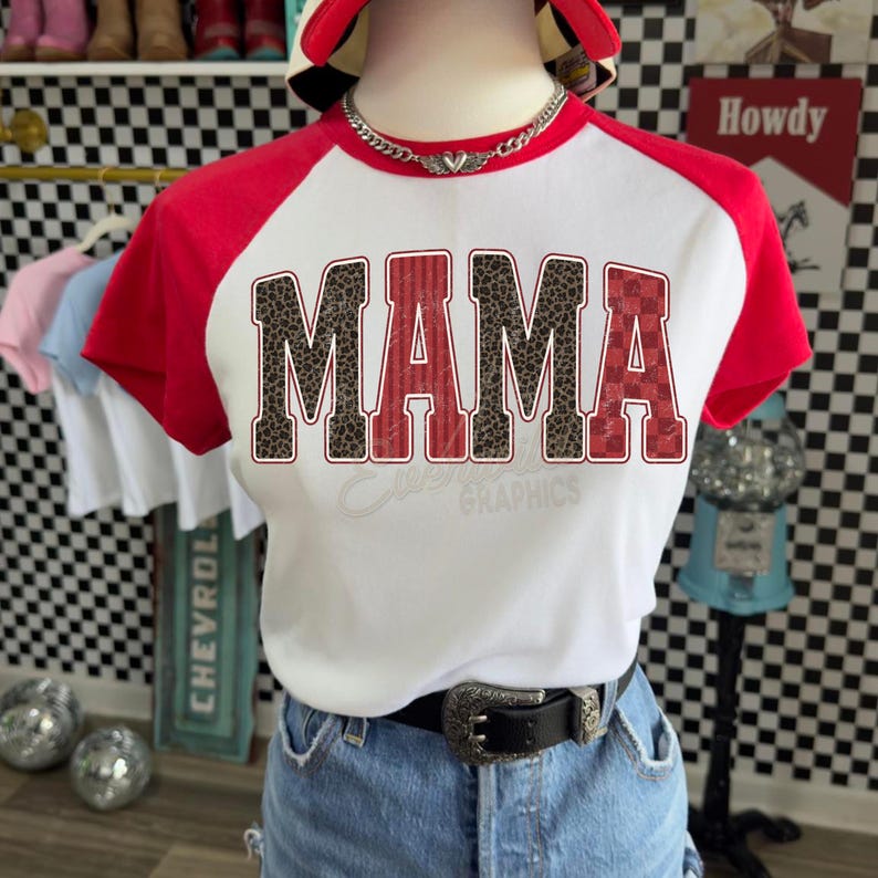 May include: A white t-shirt with red raglan sleeves featuring the word MAMA in large block letters. The letters have a brown leopard print pattern and a red checkered background. A silver chain necklace with a heart charm is worn around the neck.