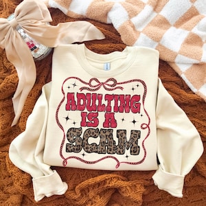 May include: Cream-coloured sweatshirt featuring the text "ADULTING IS A SCAM" in red and leopard print. The text is framed by a red decorative border with stars. The sweatshirt is displayed on a brown knitted surface.