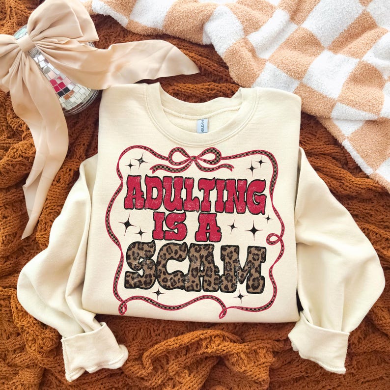 May include: Cream-coloured sweatshirt featuring the text "ADULTING IS A SCAM" in red and leopard print. The text is framed by a red decorative border with stars. The sweatshirt is displayed on a brown knitted surface.