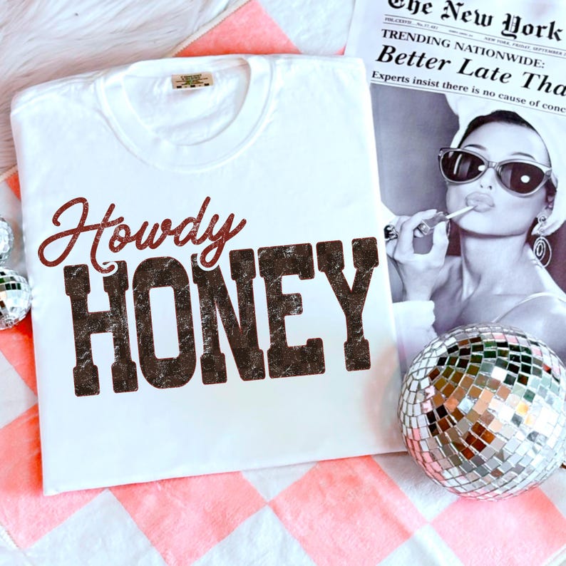May include: White t-shirt with the words "Howdy Honey" printed in a distressed brown font. The shirt is displayed on a pink and white checkered surface, with a disco ball and a vintage newspaper in the background.