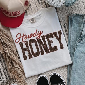 May include: A white t-shirt with the words "Howdy HONEY" in red and brown lettering. A red and beige baseball cap with the word "TIMELESS" is visible. Black and white sneakers and blue jeans are also in the image.