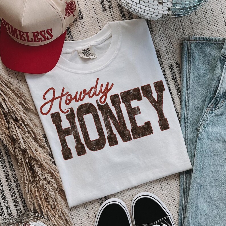 May include: A white t-shirt with the words "Howdy HONEY" in red and brown lettering. A red and beige baseball cap with the word "TIMELESS" is visible. Black and white sneakers and blue jeans are also in the image.