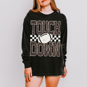 Puede incluir: Una camiseta negra de manga larga con el texto TOUCH DOWN en un patr&oacute;n de leopardo marr&oacute;n. Entre las palabras hay un gr&aacute;fico de un bal&oacute;n de f&uacute;tbol americano blanco sobre un fondo de cuadros blanco y negro. El dise&ntilde;o tiene una textura desgastada.