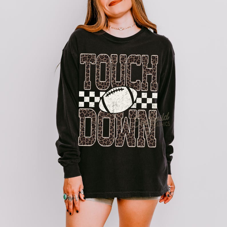 Puede incluir: Una camiseta negra de manga larga con el texto TOUCH DOWN en un patr&oacute;n de leopardo marr&oacute;n. Entre las palabras hay un gr&aacute;fico de un bal&oacute;n de f&uacute;tbol americano blanco sobre un fondo de cuadros blanco y negro. El dise&ntilde;o tiene una textura desgastada.