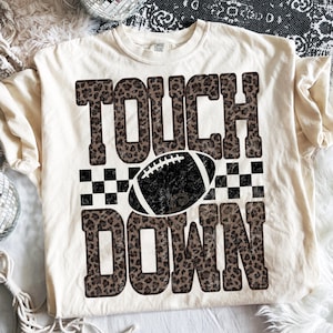 Puede incluir: Una camiseta de manga corta color crema con el texto TOUCH DOWN en un patr&oacute;n de estampado de leopardo marr&oacute;n. Entre las palabras hay un gr&aacute;fico de bal&oacute;n de f&uacute;tbol americano en blanco y negro centrado sobre un patr&oacute;n de cuadros.