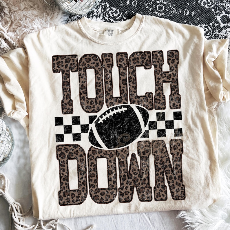 Puede incluir: Una camiseta de manga corta color crema con el texto TOUCH DOWN en un patr&oacute;n de estampado de leopardo marr&oacute;n. Entre las palabras hay un gr&aacute;fico de bal&oacute;n de f&uacute;tbol americano en blanco y negro centrado sobre un patr&oacute;n de cuadros.