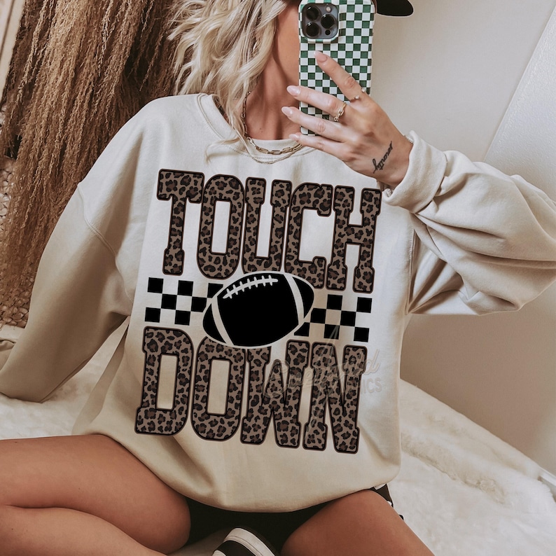 Puede incluir: Una sudadera de cuello redondo color crema con un estampado gr&aacute;fico que incluye el texto TOUCH DOWN en un patr&oacute;n de leopardo marr&oacute;n. Un icono de bal&oacute;n de f&uacute;tbol americano en blanco y negro est&aacute; centrado entre las palabras, flanqueado por cuadros.