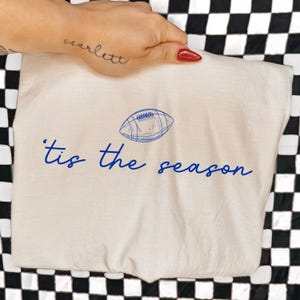 May include: A cream-colored t-shirt featuring a blue line-art illustration of an American football centered above the blue cursive text 'tis the season'. The design is printed on the front of the fabric, showcasing a simple and clean graphic style.