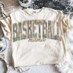 May include: A cream-coloured t-shirt featuring the words "BASKETBALL" and "mama". The text is in a brown, collegiate-style font, with "BASKETBALL" filled with a camouflage pattern. The shirt is made of a soft material and is suitable for casual wear.