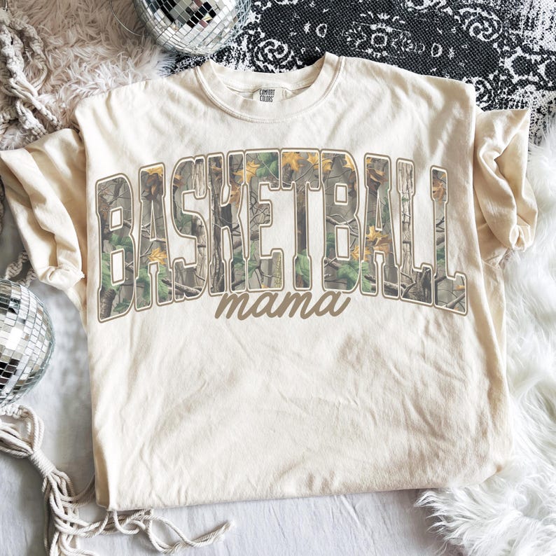 May include: A cream-coloured t-shirt featuring the words "BASKETBALL" and "mama". The text is in a brown, collegiate-style font, with "BASKETBALL" filled with a camouflage pattern. The shirt is made of a soft material and is suitable for casual wear.