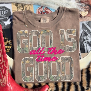 K&ouml;nnte beinhalten: Ein braunes Sweatshirt mit dem Text "GOD IS all the GOOD" in einem Camouflage-Muster und rosa Schriftzug. Das Sweatshirt ist gefaltet und hat ein Comfort Colors-Etikett. Eine goldene Kreuzkette liegt obenauf.
