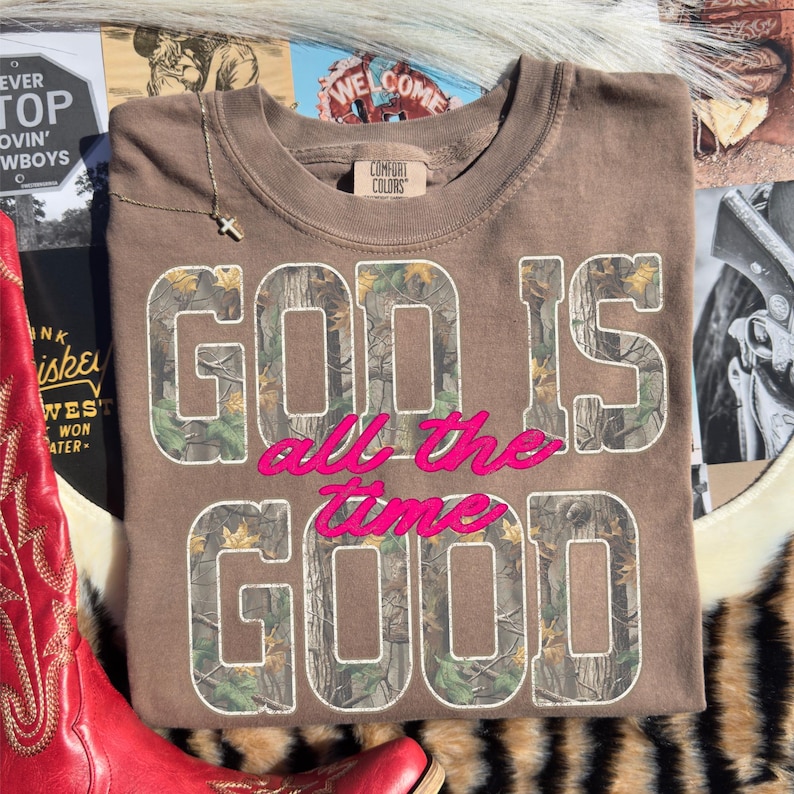 K&ouml;nnte beinhalten: Ein braunes Sweatshirt mit dem Text "GOD IS all the GOOD" in einem Camouflage-Muster und rosa Schriftzug. Das Sweatshirt ist gefaltet und hat ein Comfort Colors-Etikett. Eine goldene Kreuzkette liegt obenauf.