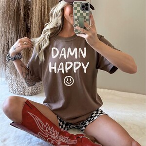 May include: A brown t-shirt with the words "DAMN HAPPY" and a smiley face graphic. The person is wearing a brown and tan trucker hat, black and white checkered shorts, and red cowboy boots.