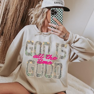 K&ouml;nnte beinhalten: Beiges Sweatshirt mit dem Text "GOD IS all the time GOOD" in einem Camouflage-Muster und rosa Schriftzug. Die Person tr&auml;gt eine wei&szlig;e M&uuml;tze mit schwarzem Text und eine gr&uuml;n-wei&szlig; karierte Handyh&uuml;lle.