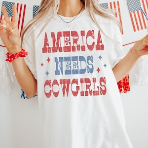 Może przedstawiać: Biała koszulka z napisem "AMERICA NEEDS COWGIRLS" w kolorze czerwonym i niebieskim. Projekt ma wygląd vintage i postarzaną stylistykę. Patriotyczny motyw koszulki sprawia, że nadaje się do noszenia na co dzień. Projekt zawiera gwiazdy.