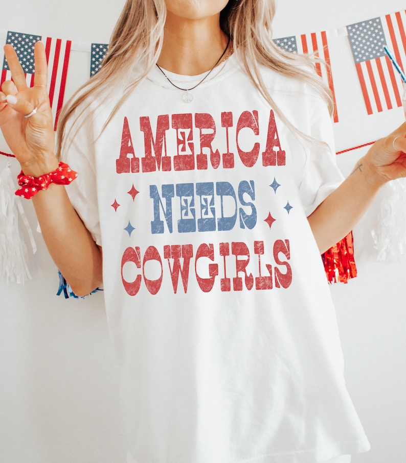Może przedstawiać: Biała koszulka z napisem "AMERICA NEEDS COWGIRLS" w kolorze czerwonym i niebieskim. Projekt ma wygląd vintage i postarzaną stylistykę. Patriotyczny motyw koszulki sprawia, że nadaje się do noszenia na co dzień. Projekt zawiera gwiazdy.