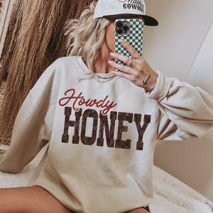 May include: Cream-colored sweatshirt with the words "Howdy HONEY" in red and brown lettering. The person is wearing a white baseball cap with black text and a green and white checkered phone case.