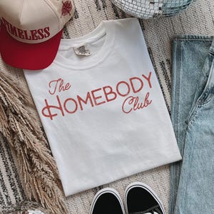 K&ouml;nnte beinhalten: Ein wei&szlig;es T-Shirt mit dem Text "The Homebody Club" in roter Schrift. Eine rote und beige Baseballkappe mit dem Wort "TIMELESS" ist ebenfalls zu sehen. Schwarz-wei&szlig;e Turnschuhe und blaue Jeans sind ebenfalls im Bild.