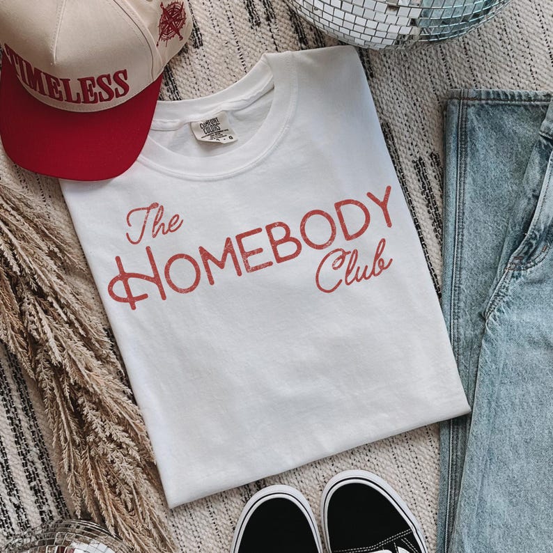K&ouml;nnte beinhalten: Ein wei&szlig;es T-Shirt mit dem Text "The Homebody Club" in roter Schrift. Eine rote und beige Baseballkappe mit dem Wort "TIMELESS" ist ebenfalls zu sehen. Schwarz-wei&szlig;e Turnschuhe und blaue Jeans sind ebenfalls im Bild.