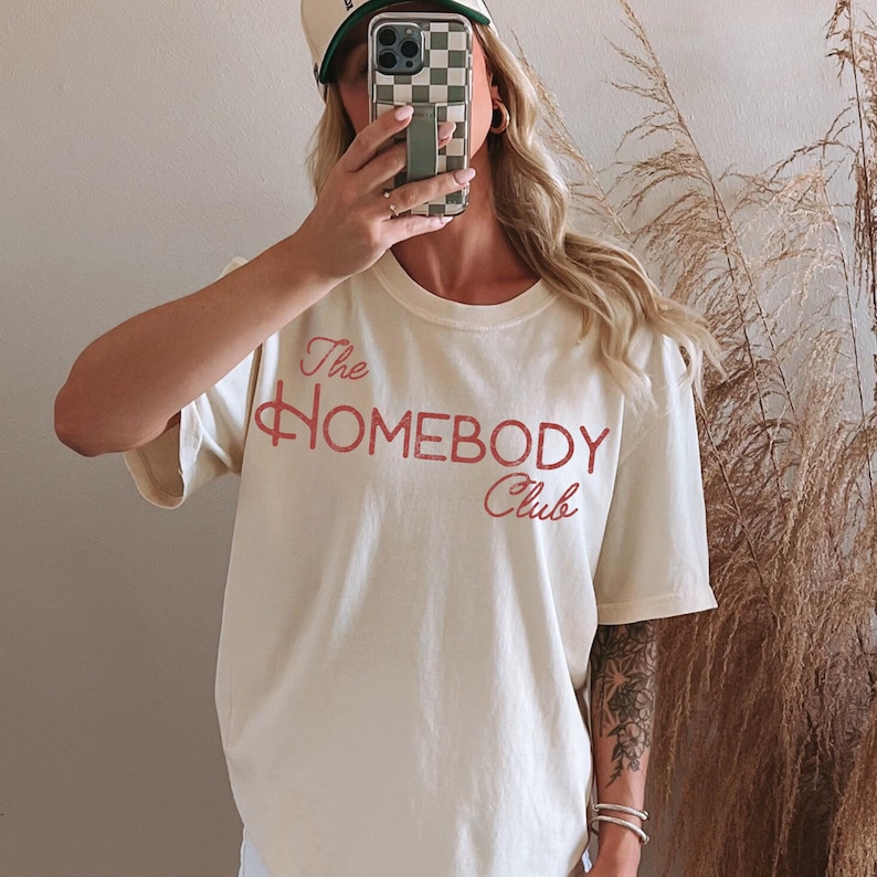 K&ouml;nnte beinhalten: Cremefarbenes T-Shirt mit dem Text "The Homebody Club" in roter Schrift. Das Shirt hat kurze &Auml;rmel und einen Rundhalsausschnitt. Eine Baseballkappe mit Schachbrettmuster ist ebenfalls zu sehen.