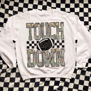 May include: A white crewneck sweatshirt featuring the text TOUCH DOWN in large block letters with a camouflage pattern. A black and white football graphic is positioned between the words, flanked by a black and white checkered pattern.