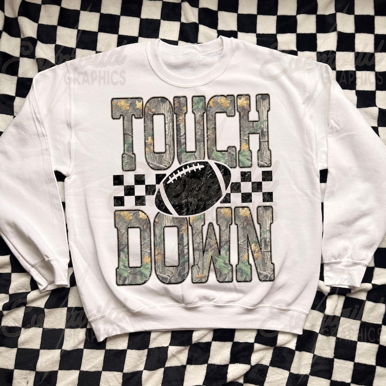 May include: A white crewneck sweatshirt featuring the text TOUCH DOWN in large block letters with a camouflage pattern. A black and white football graphic is positioned between the words, flanked by a black and white checkered pattern.