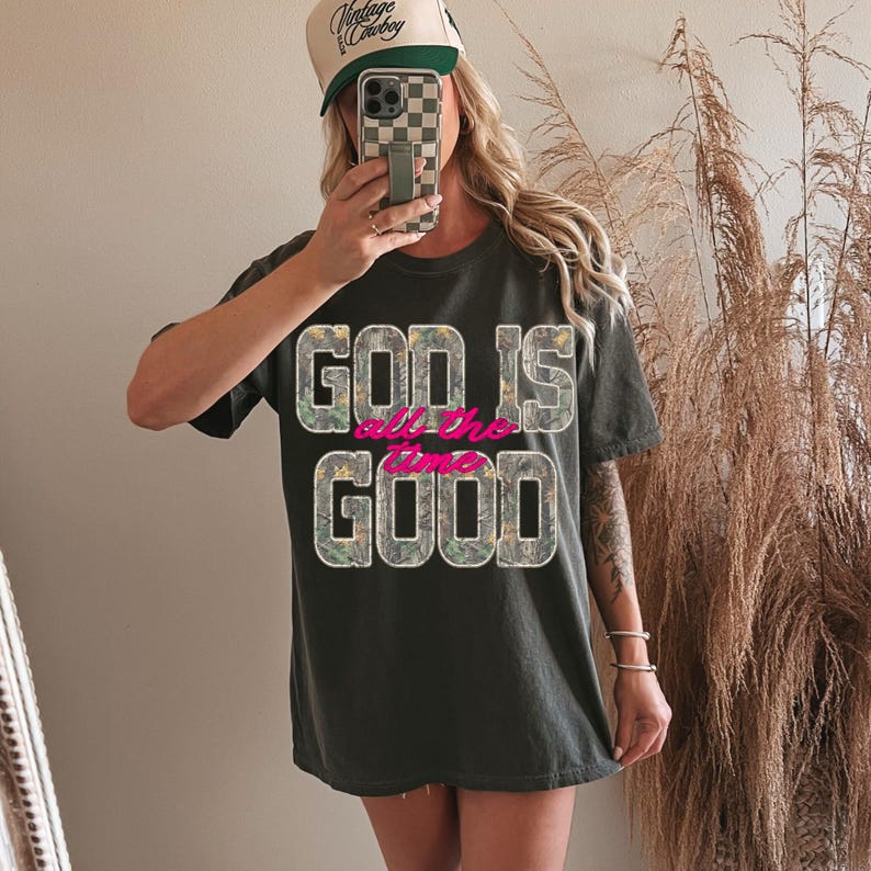 K&ouml;nnte beinhalten: Dunkelgraues T-Shirt mit dem Text "GOD IS all the time GOOD" in Tarnfarben und Pink. Die Person tr&auml;gt eine beige-gr&uuml;ne Trucker-M&uuml;tze mit dem Schriftzug "Vintage Cowboy".