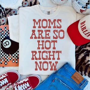 May include: A white t-shirt with the text "MOMS ARE SO HOT RIGHT NOW" in red letters. The image also includes a red and white baseball cap, blue jeans, and red sneakers.