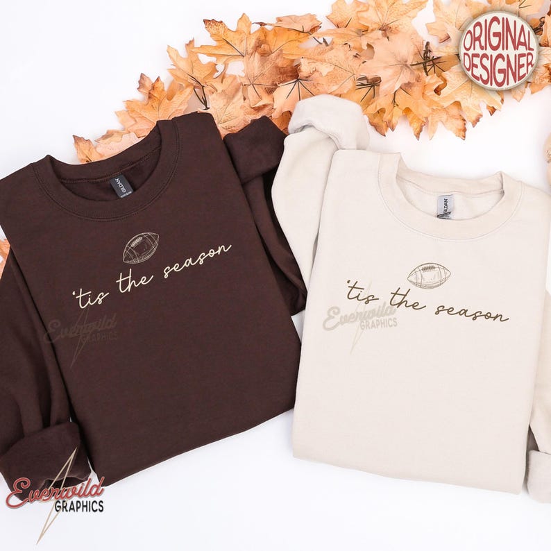 May include: A dark brown sweatshirt and a cream-colored sweatshirt, both featuring a graphic of a football and the text 'tis the season in a cursive font. The garments have crew necklines and long sleeves, presented as a pair of matching tops.