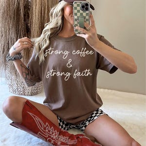 May include: A brown t-shirt with the text "strong coffee & strong faith" in white script. The person is wearing a brown and tan trucker hat, black and white checkered shorts, and red cowboy boots.