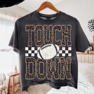 Puede incluir: Una camiseta de manga corta gris carb&oacute;n con el texto TOUCH DOWN en un patr&oacute;n de estampado de leopardo marr&oacute;n y negro. Un gr&aacute;fico de bal&oacute;n de f&uacute;tbol americano blanco con una franja a cuadros blancos y negros se sit&uacute;a entre las palabras.
