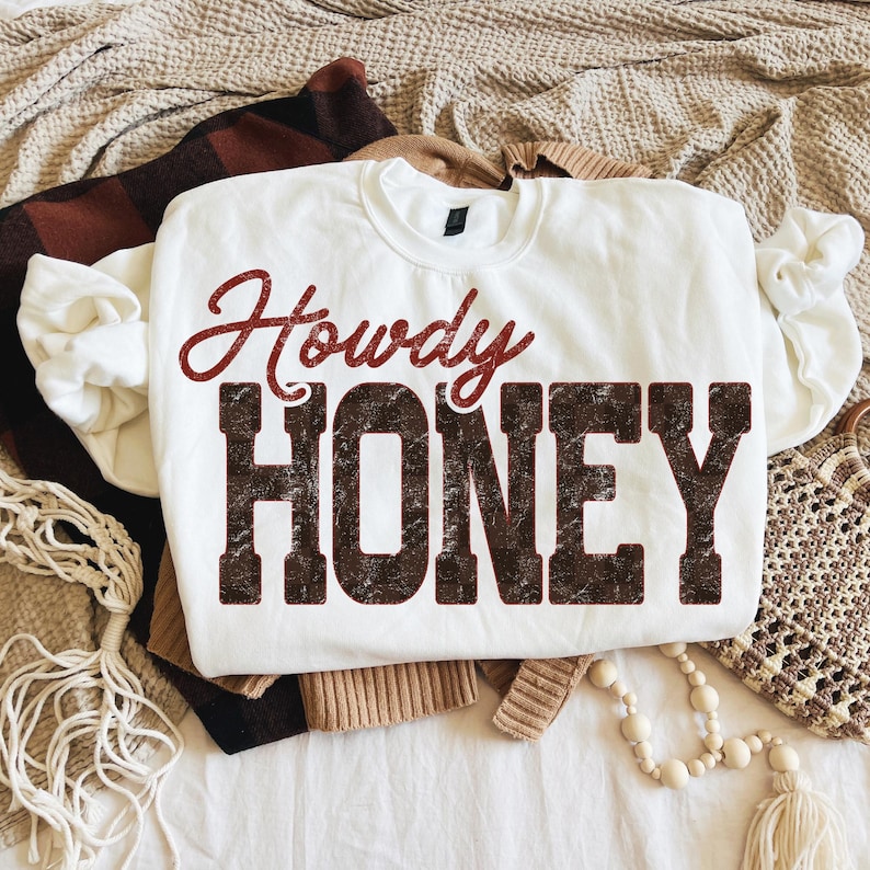 May include: White crewneck sweatshirt with the words "Howdy HONEY" printed in a distressed, brown font. The word "Howdy" is in a cursive, red font. The sweatshirt is folded on top of other clothing items.