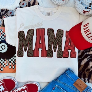 May include: A white short-sleeve t-shirt featuring the word MAMA in large block letters. The letters have varied patterns including brown leopard print, red vertical stripes, and a red checkered pattern. The text Everwild Graphics is visible.