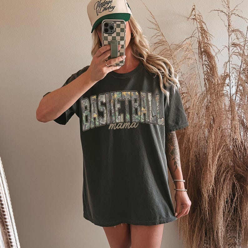 May include: Dark grey t-shirt with the text "BASKETBALL mama" in a camouflage design. The person in the image is wearing a trucker hat with the text "Vintage Cowboy". The hat is white and green.