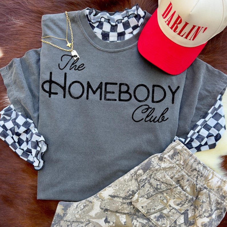 K&ouml;nnte beinhalten: Ein graues T-Shirt mit dem Text "The Homebody Club" in schwarzer Schrift, &uuml;berlagert von einem lang&auml;rmeligen Oberteil mit schwarz-wei&szlig;em Schachbrettmuster. Eine rote und beige Baseballkappe mit "Darlin'" in roter Schrift liegt daneben, zusammen mit einer goldenen Halskette und Camouflage-Hosen.